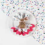 Confetti-patterned cloth napkin styled on a white plate with a woven bunny napkin ring and pink scalloped trim.