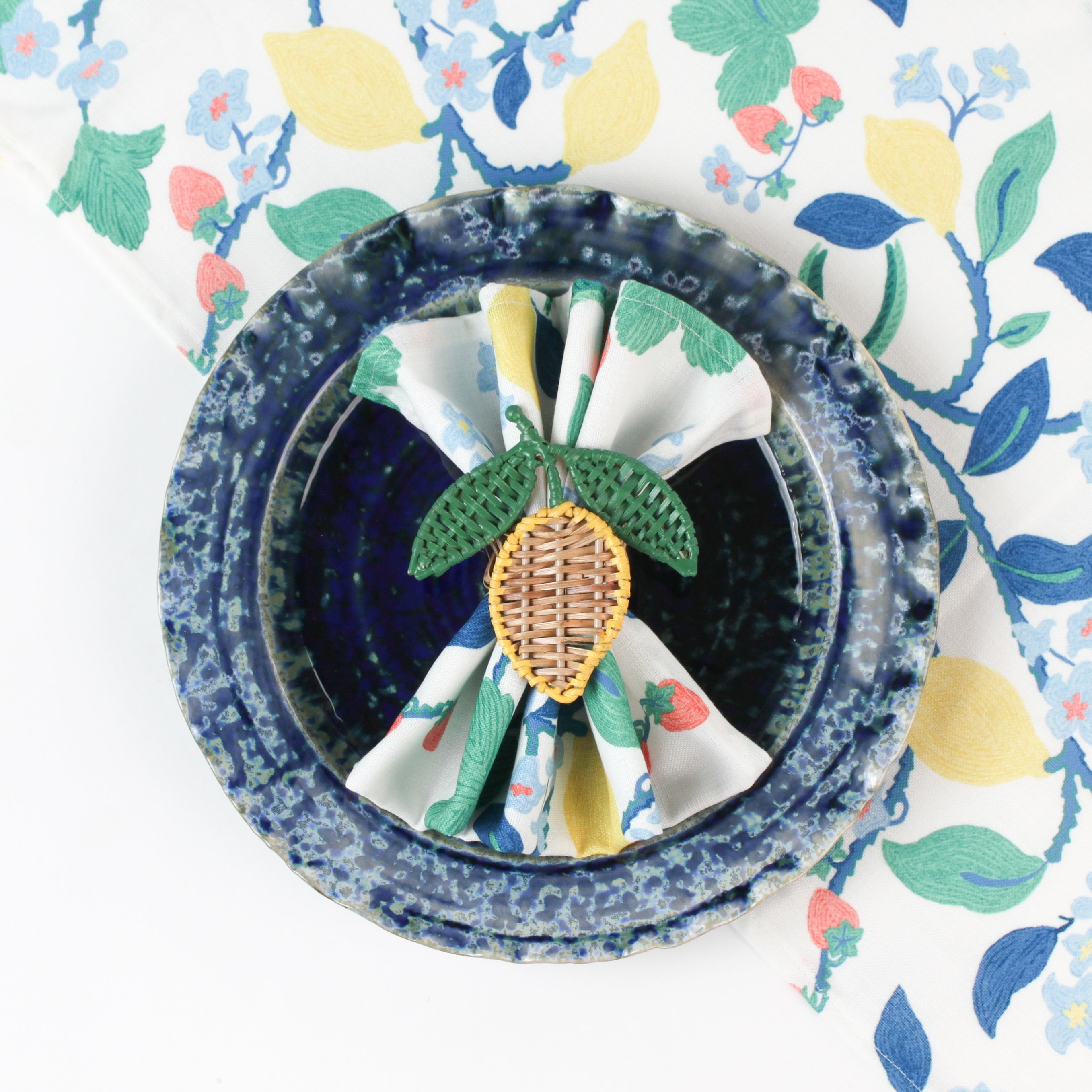 Botanical print cloth napkin placed in a blue textured bowl with a woven lemon napkin ring.