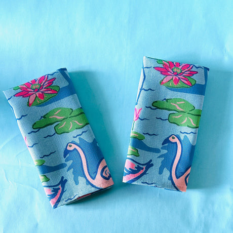 A folded pair of Sunken Gardens Flamingo cloth dinner napkins on a turquoise background