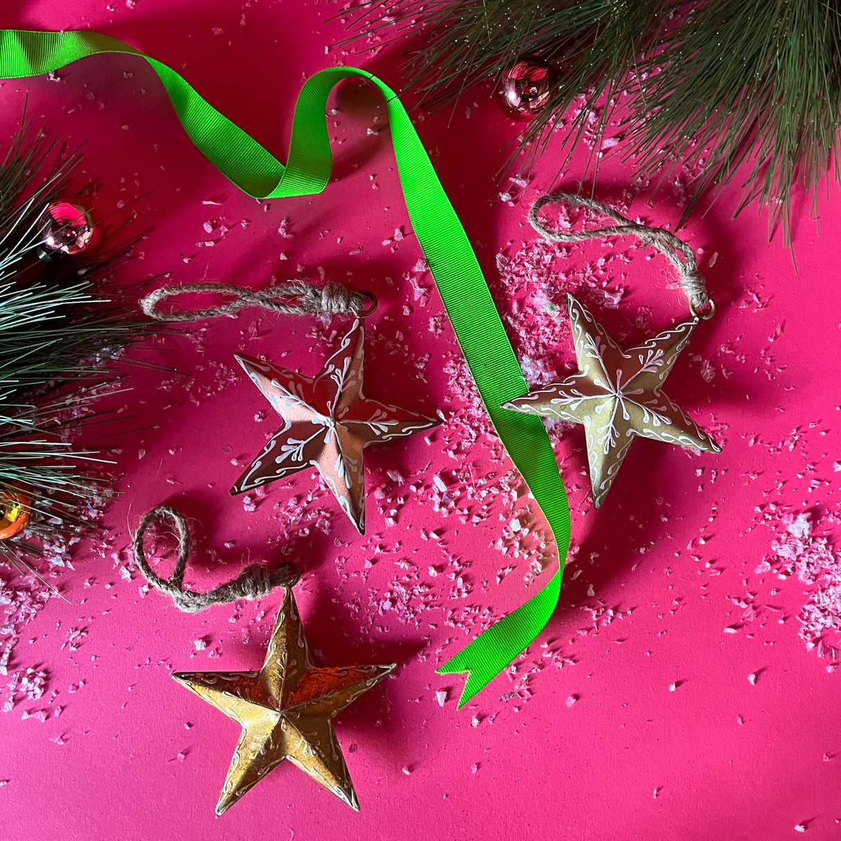 Three star christmas ornaments that feature white painted embellishments. Resting on a red background with snow, green ribbon and pine leaves.