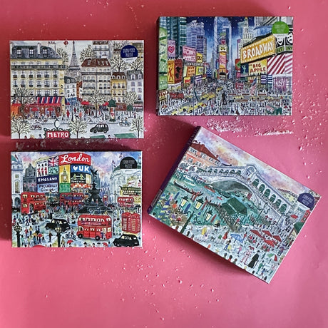 Assorted travel-themed jigsaw puzzles depicting Times Square, London, Venice, and Paris, displayed on a pink background.
