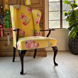 A vintage wingback chair with yellow upholstery featuring a floral pattern, positioned in a room with wooden flooring and a plant in the background.