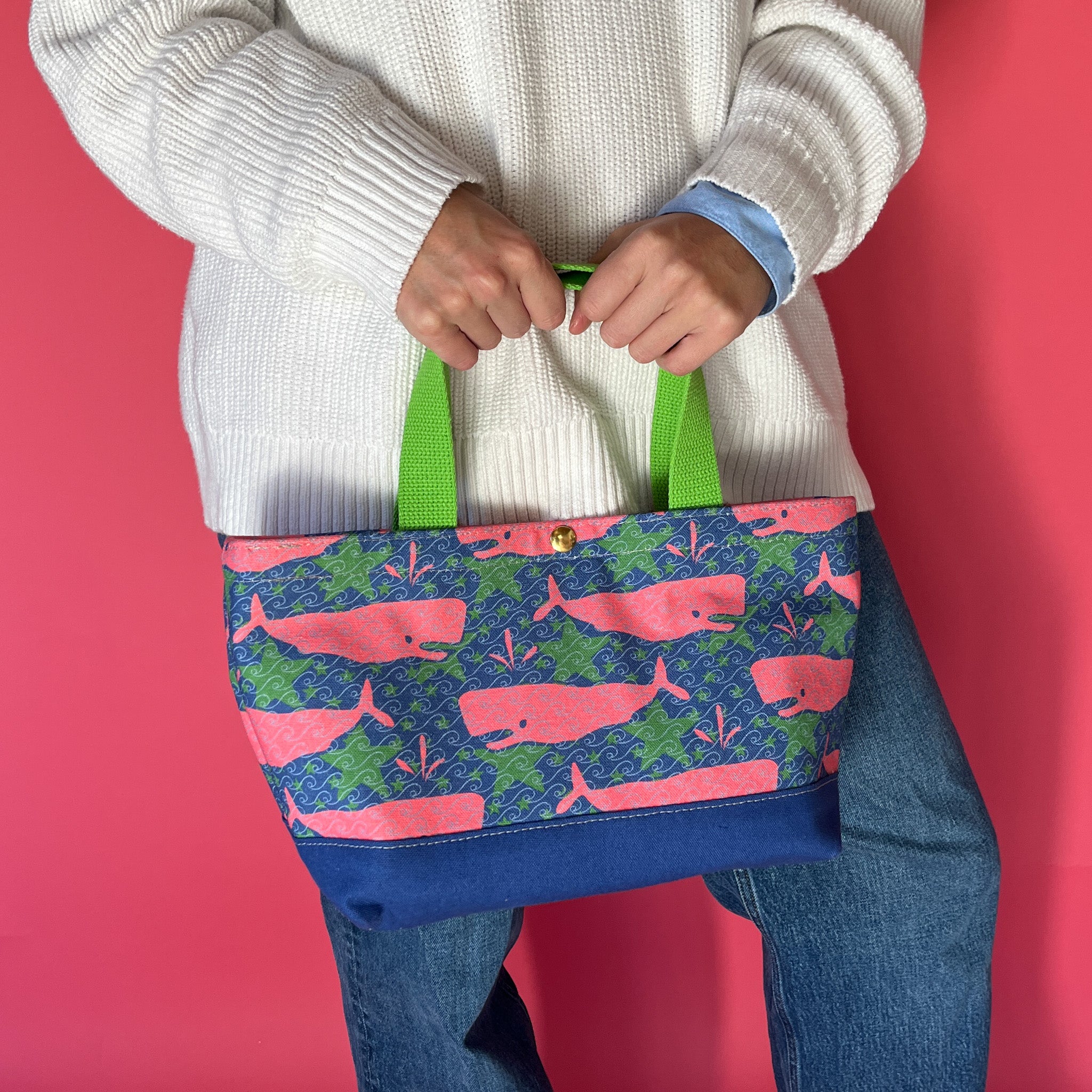 Whale of a Time canvas small tote bag with lime straps and blue fabric with a repeating pink whale  and green star motif being held by model.