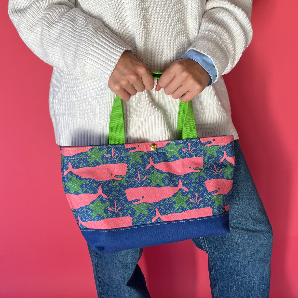 Whale of a Time canvas small tote bag with lime straps and blue fabric with a repeating pink whale  and green star motif being held by model.