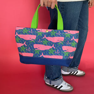 Whale of a Time small canvas tote bag with lime straps and blue fabric with a repeating pink whale  and green star motif hanging from models hand.