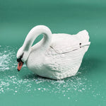 White ceramic swan-shaped soup tureen with lid and ladle on a green background with faux snow