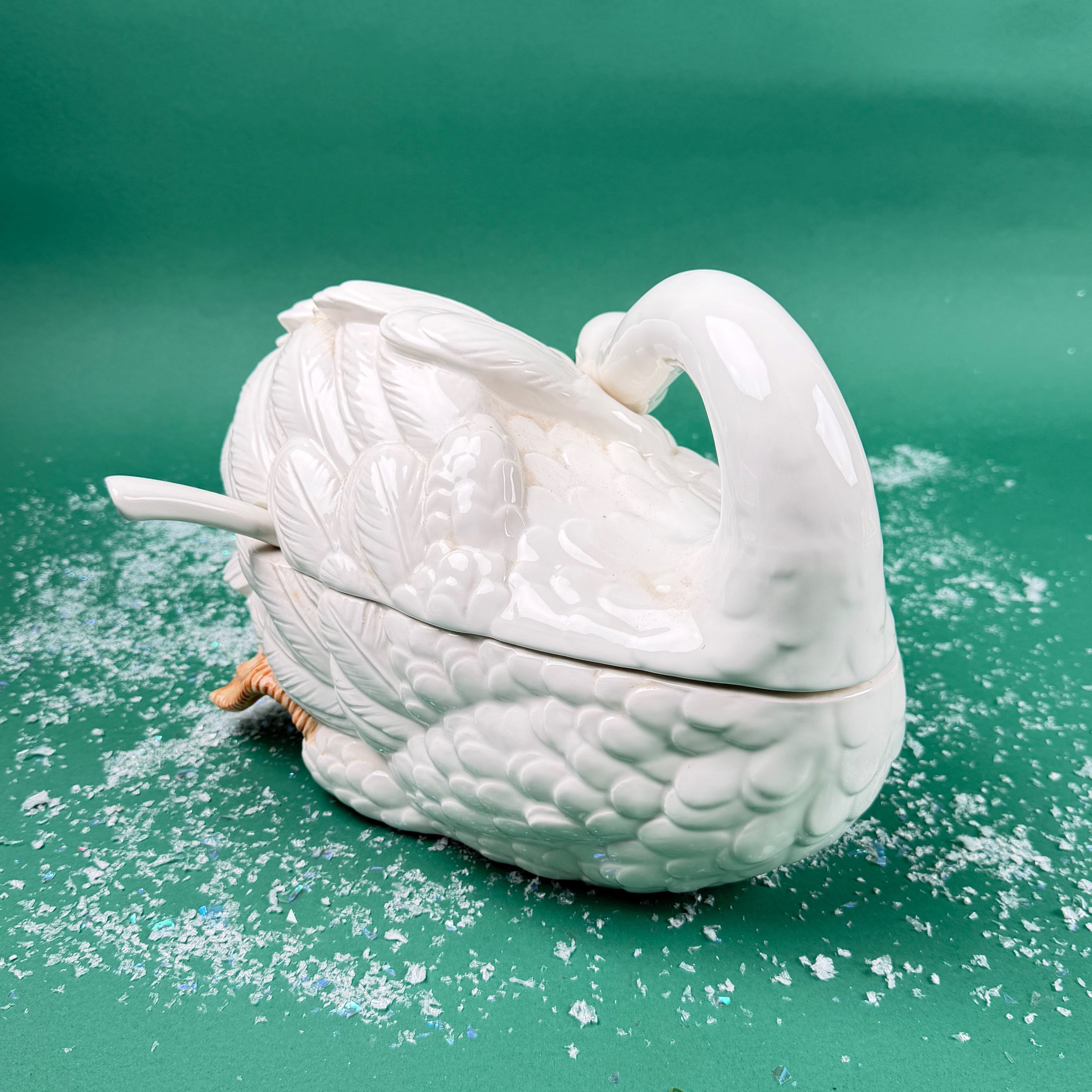 Side view of a white ceramic swan-shaped soup tureen with ladle on a green background with faux snow