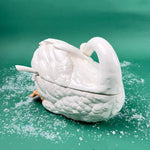 Side view of a white ceramic swan-shaped soup tureen with ladle on a green background with faux snow