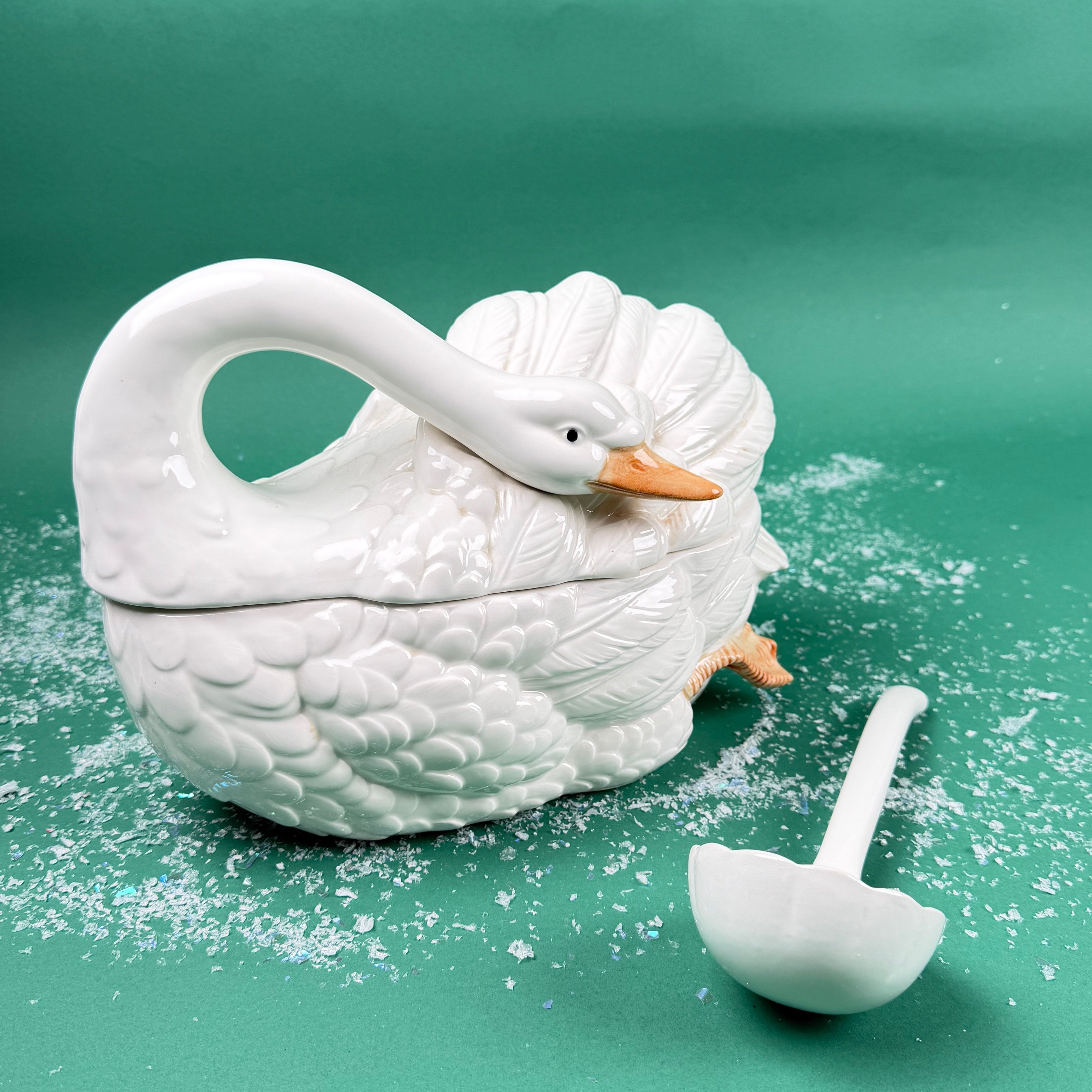White ceramic swan-shaped soup tureen with lid and separate ladle on a green background with faux snow