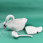 Open white ceramic swan soup tureen with detached lid and matching ladle on a green background with faux snow