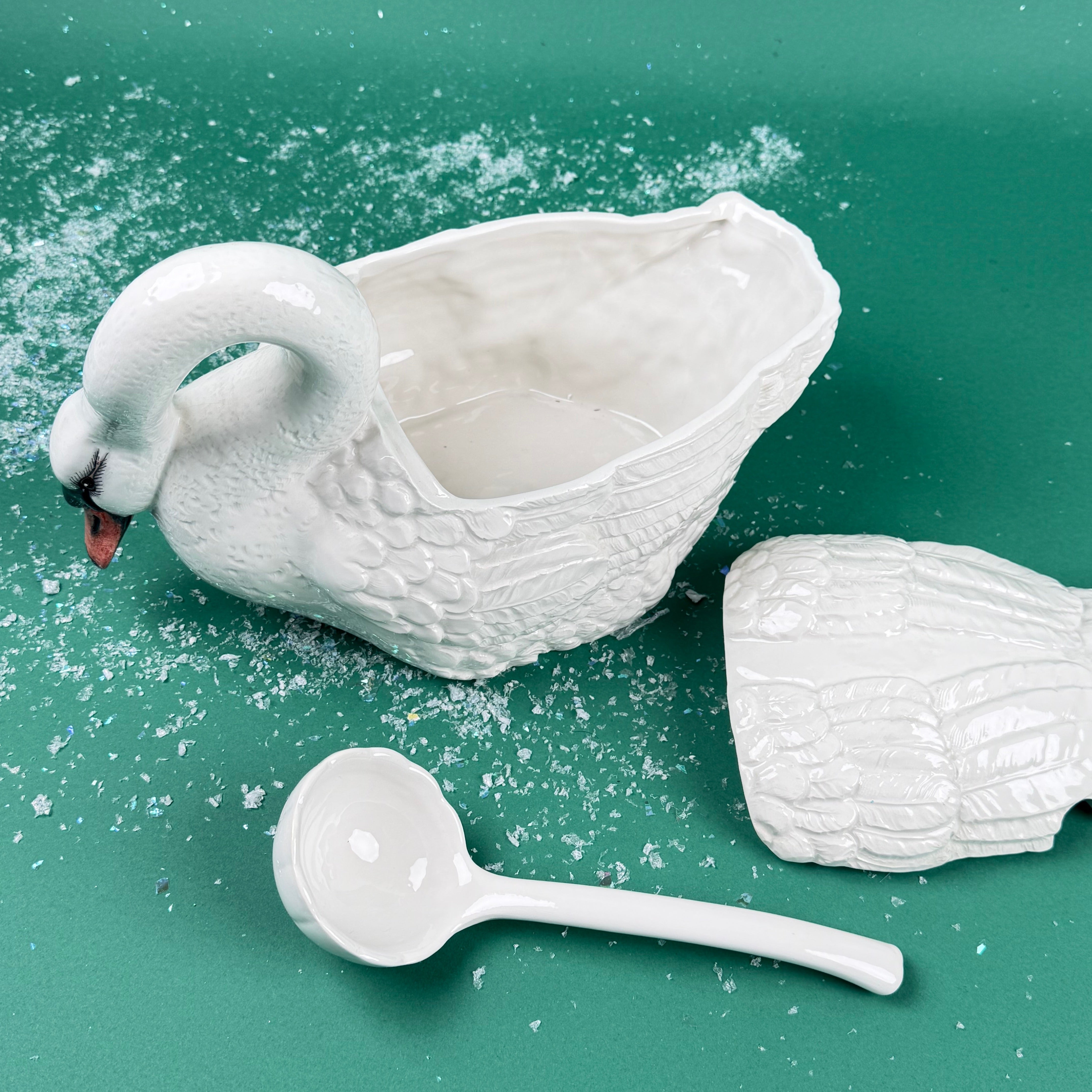 Open white ceramic swan soup tureen with detached lid and matching ladle on a green background with faux snow