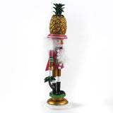 side view of a 19.5" tall Yellow and Pink Christmas Nutcracker Table Decoration with a gold pineapple hat and  holding a palm tree against a white background. 