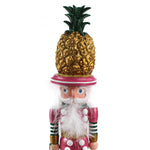 close up of a 19.5" tall Yellow and Pink Christmas Nutcracker Table Decoration with a gold pineapple hat and  holding a palm tree against a white background. 