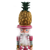 close up of a 19.5" tall Yellow and Pink Christmas Nutcracker Table Decoration with a gold pineapple hat and  holding a palm tree against a white background. 