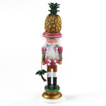a 19.5" tall Yellow and Pink Christmas Nutcracker Table Decoration with a gold pineapple hat and  holding a palm tree against a white background. 