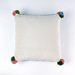 back of a Square hooked wool christmas pillow featuring a green Christmas ornament with red, pink, and blue patterns on a white background with a light blue scalloped border and colorful pom-poms on each corner.