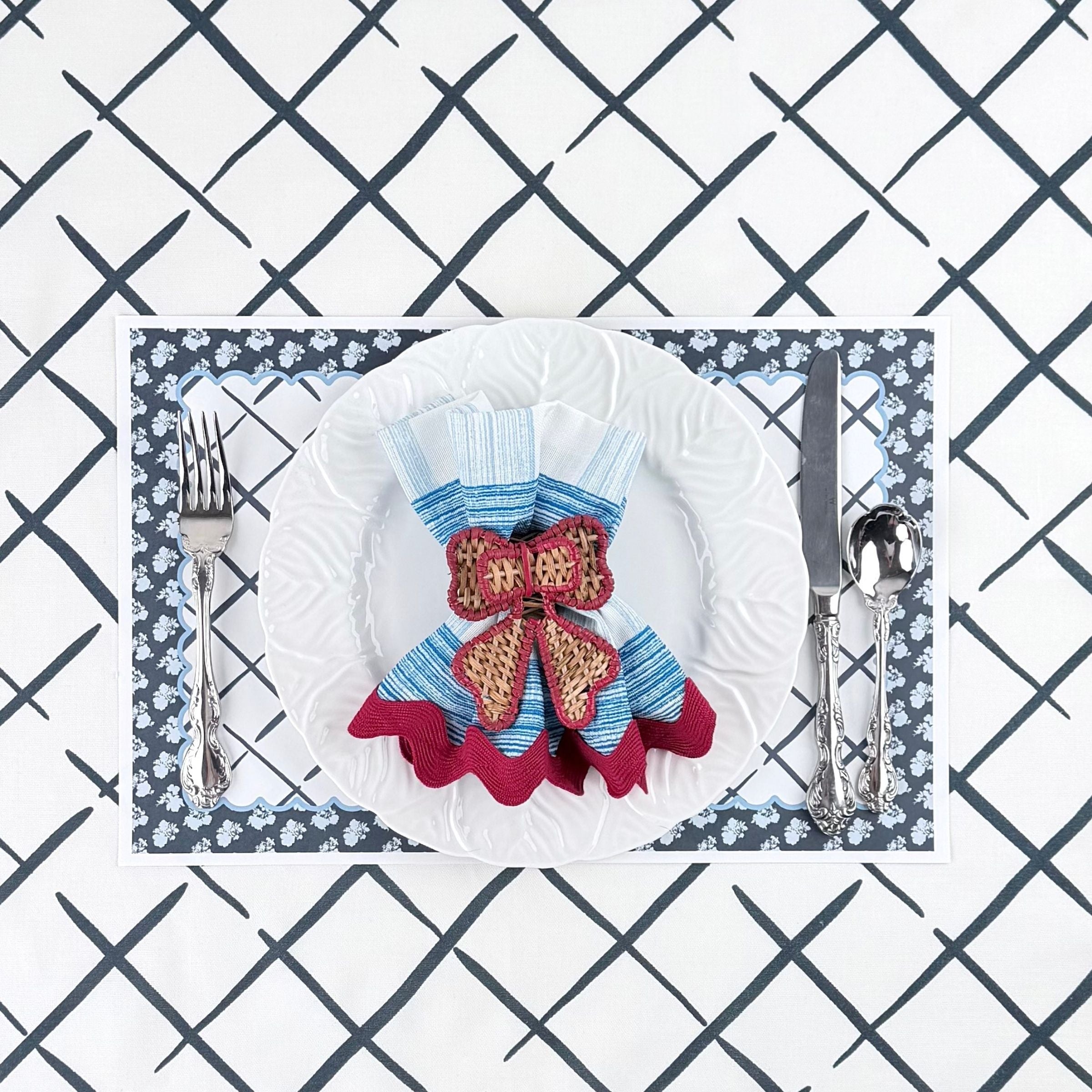 a decorative place setting with a bow-shaped napkin on a white plate on top of a blue and white floral paper placemat with a geometric background.