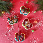 A set of four beaded butterfly felt Christmas ornaments in red, green, blue, and yellow colors, displayed against a pink background with a sprinkling of snowflakes and decorative branches.