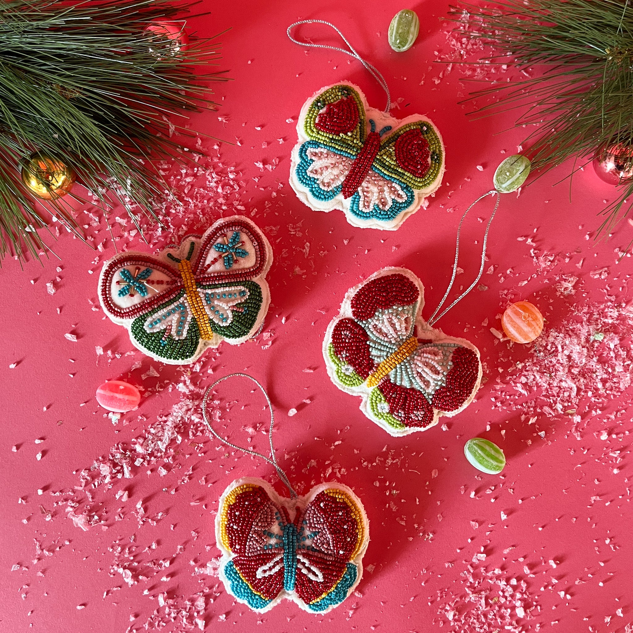 A set of four beaded butterfly felt Christmas ornaments in red, green, blue, and yellow colors, displayed against a pink background with a sprinkling of snowflakes and decorative branches.