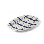 Oval ceramic serving platter with hand-painted blue plaid pattern on a white background viewed at an angled side perspective.