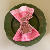 a pink gingham dinner napkin in gin lane print fabric by madap cottage with a rattan bunny napkin ring resting on a green plate