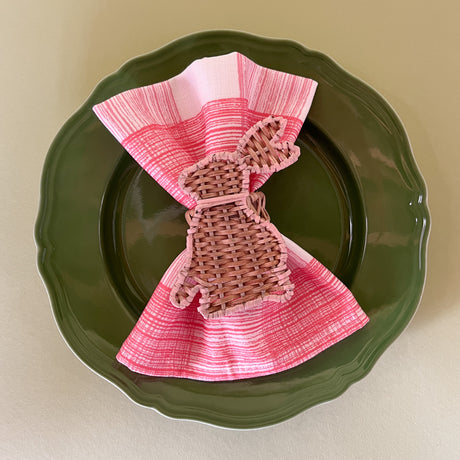 a pink gingham dinner napkin in gin lane print fabric by madap cottage with a rattan bunny napkin ring resting on a green plate