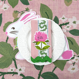 A white easter bunny placemat topped with a white plate and floral napkin and flower place card against a pink floral tablecloth.