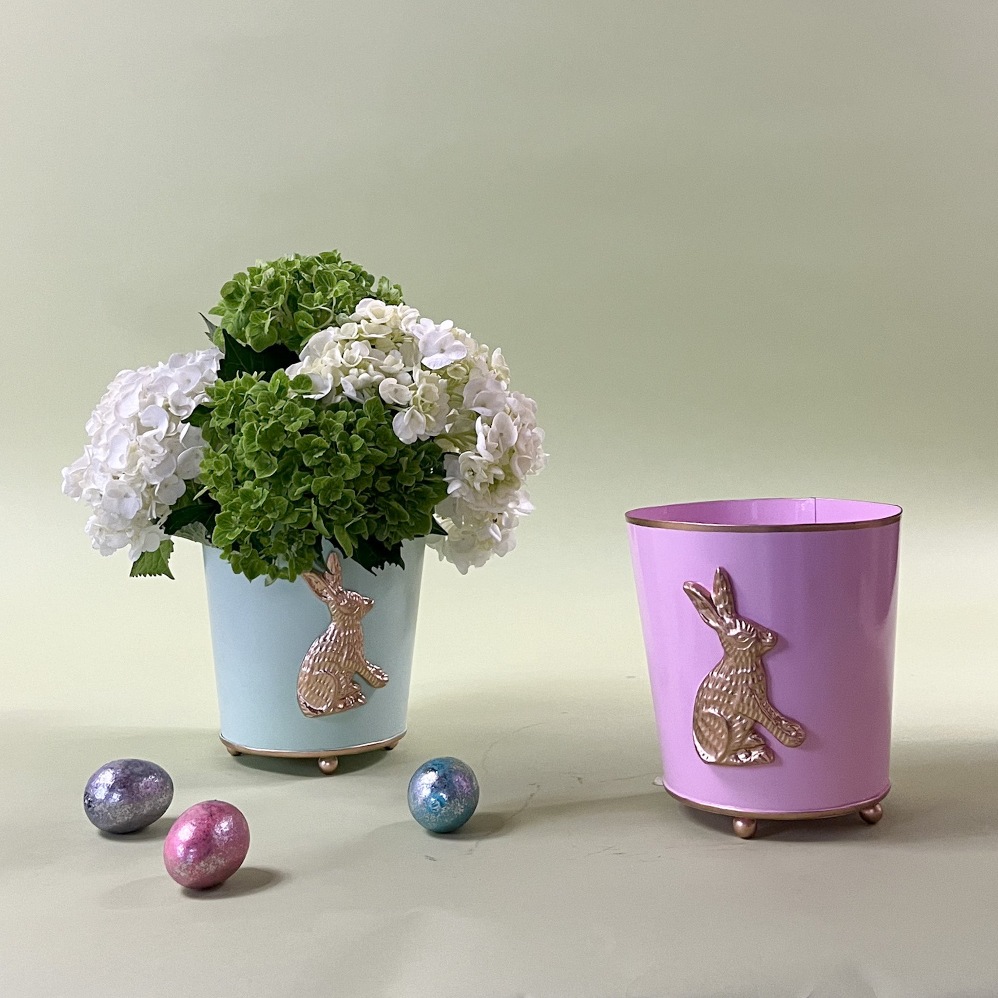 a green tole cachepot with a gold bunny motif and a pink tole cachepot with a bunny motif on a green background.