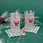 Two holiday tumblers with red candy cane bow design filled with a clear drink, placed on patterned coasters with faux snow and Christmas décor