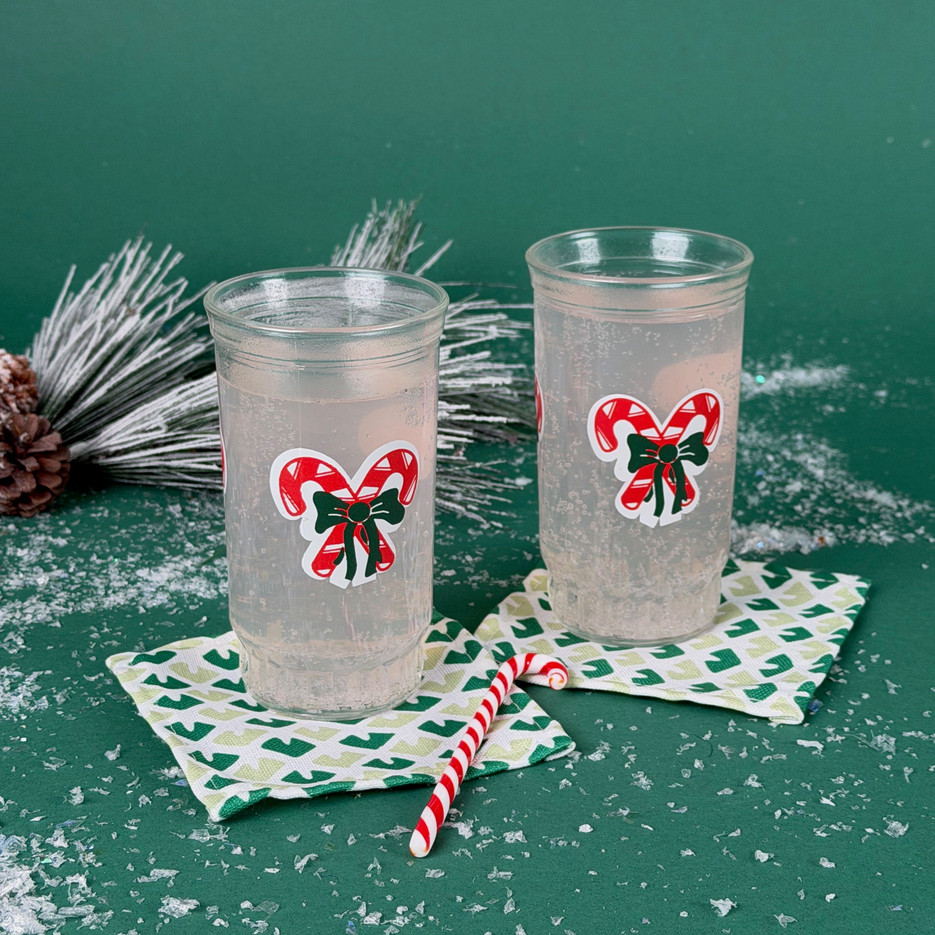 Two holiday tumblers with red candy cane bow design filled with a clear drink, placed on patterned coasters with faux snow and Christmas décor