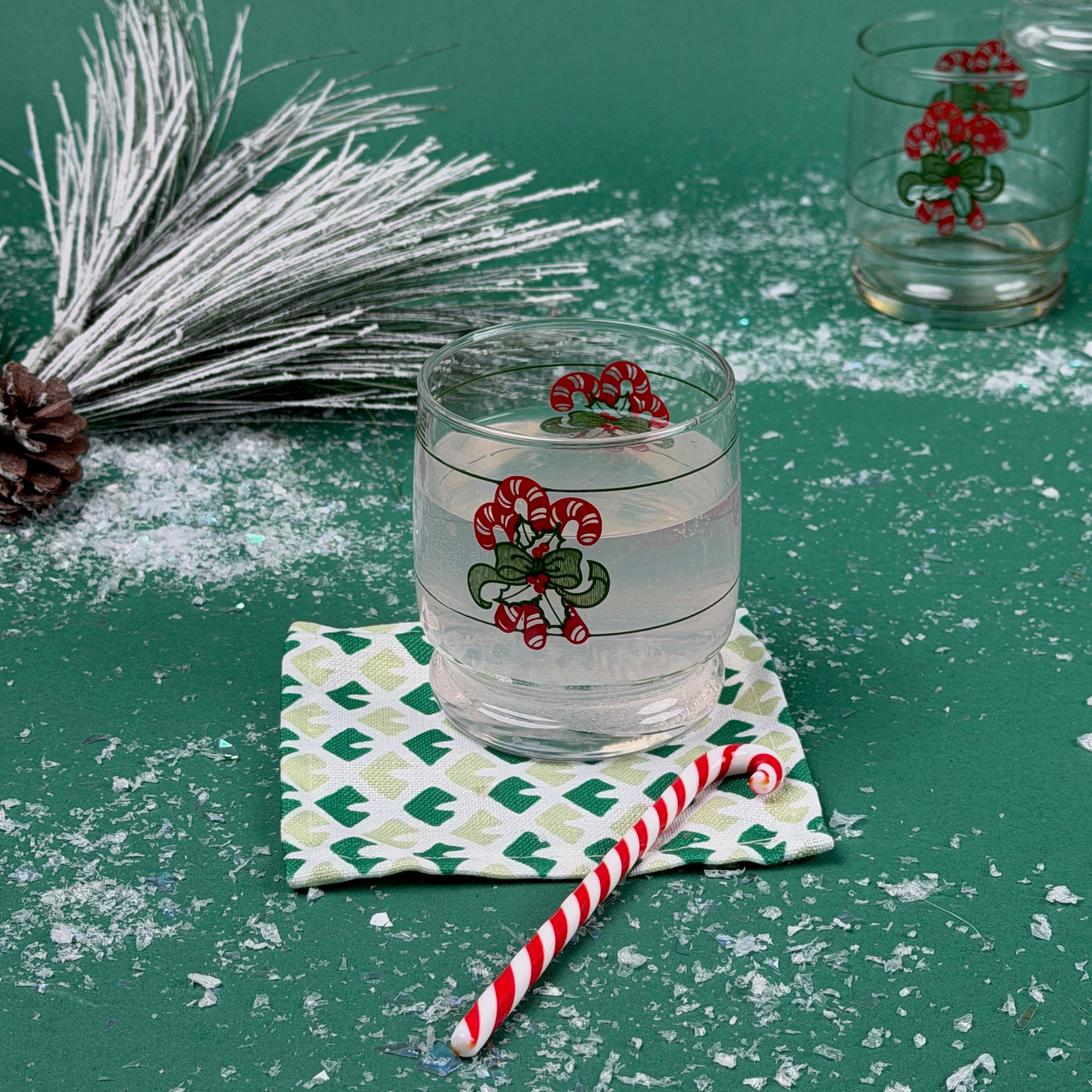 Holiday glass with a candy cane design placed on a patterned coaster with faux snow and Christmas décor on a green background