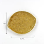 Christmas tea towel styled product photo of an oval stoneware platter in a warm ochre yellow glaze, featuring an organic uneven rim, raised textured surface with small dot detailing, shown with visible dimensions of approximately 10.375 inches wide by 8.25 inches tall on a white background.