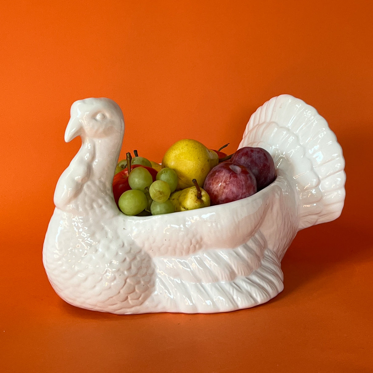 A white ceramic turkey centerpiece bowl shaped like a turkey, filled with various fruits and vegetables, against an orange background.