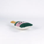 Bottle-shaped decorative pillow with a hooked texture, designed like a champagne bottle in green, pink, and gold colors, featuring the word “Cheers” on the label lying down a white background