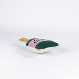 Bottle-shaped decorative pillow with a hooked texture, designed like a champagne bottle in green, pink, and gold colors, featuring the word “Cheers” on the label lying down a white background