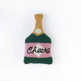 Bottle-shaped decorative pillow with a hooked texture, designed like a champagne bottle in green, pink, and gold colors, featuring the word “Cheers” on the label.
