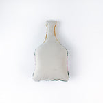 back of a bottle-shaped decorative pillow with a hooked texture, designed like a champagne bottle in green, pink, and gold colors, featuring the word “Cheers” on the label.