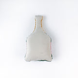 back of a bottle-shaped decorative pillow with a hooked texture, designed like a champagne bottle in green, pink, and gold colors, featuring the word “Cheers” on the label.