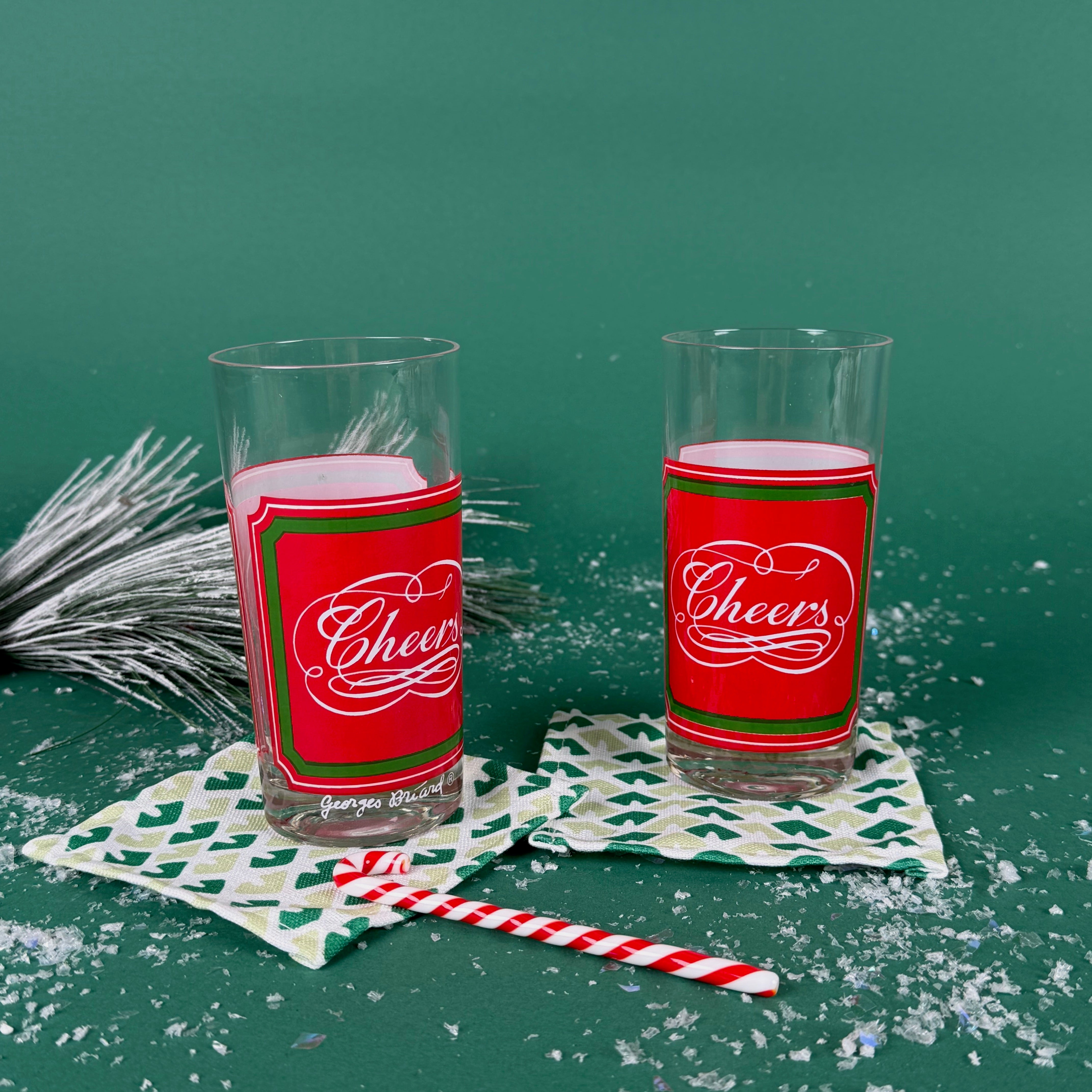 Two clear drinking glasses with red and green “Cheers” holiday labels on patterned coasters with faux snow and festive décor
