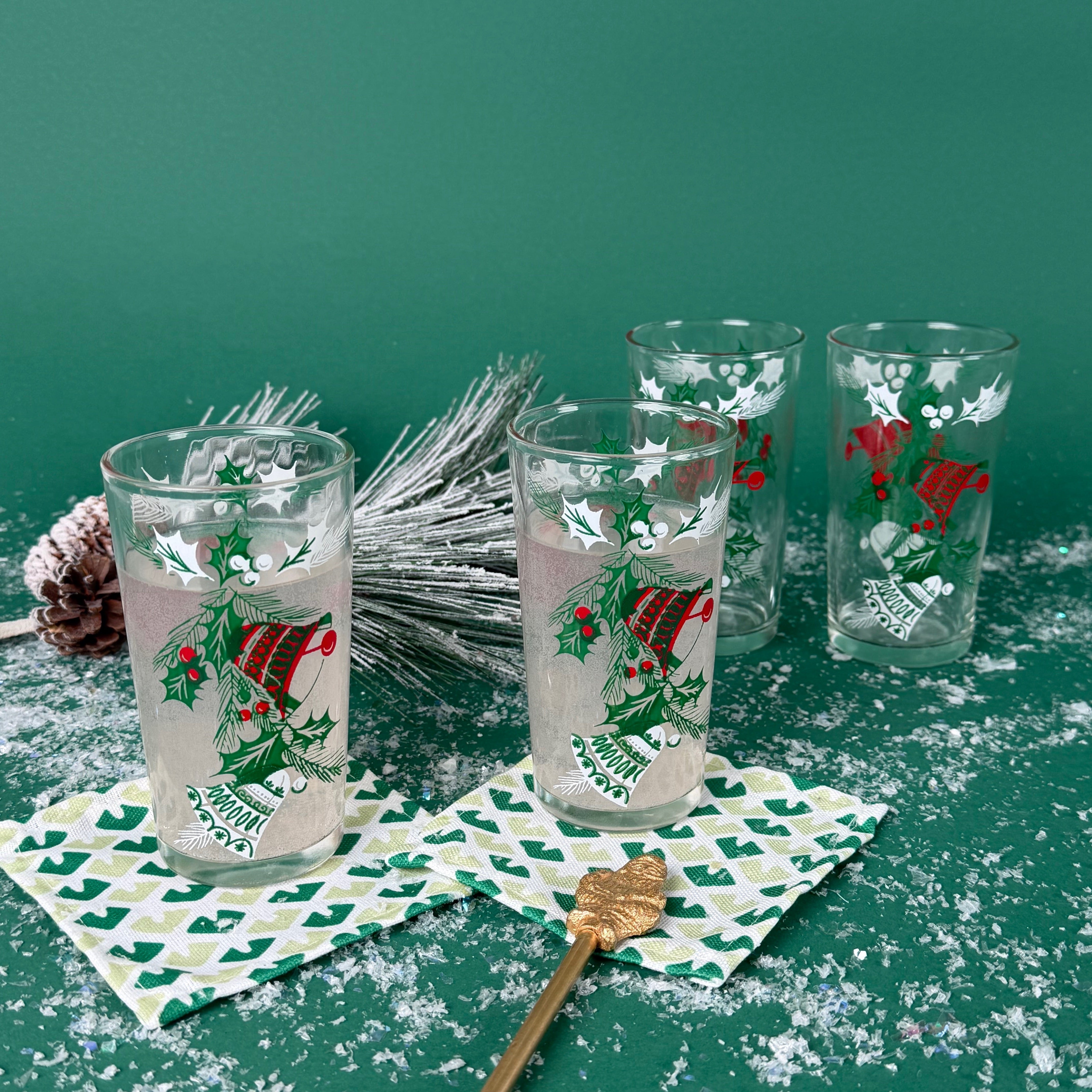Set of Christmas glasses with holly and bell designs, displayed with holiday décor and faux snow on a green background