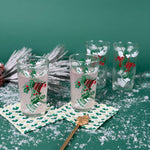 Set of Christmas glasses with holly and bell designs, displayed with holiday décor and faux snow on a green background