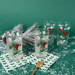 Set of Christmas glasses with holly and bell designs, displayed with holiday décor and faux snow on a green background