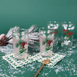 Set of Christmas glasses with holly and bell designs, displayed with holiday décor and faux snow on a green background
