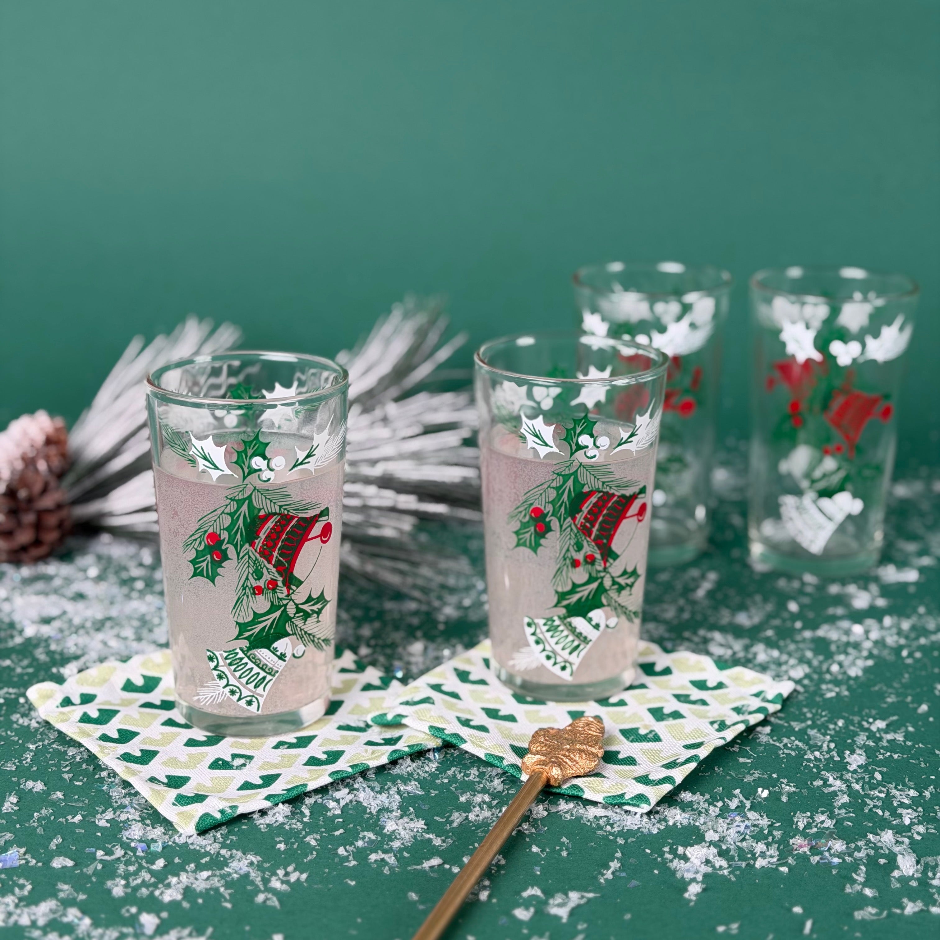Set of Christmas glasses with holly and bell designs, displayed with holiday décor and faux snow on a green background