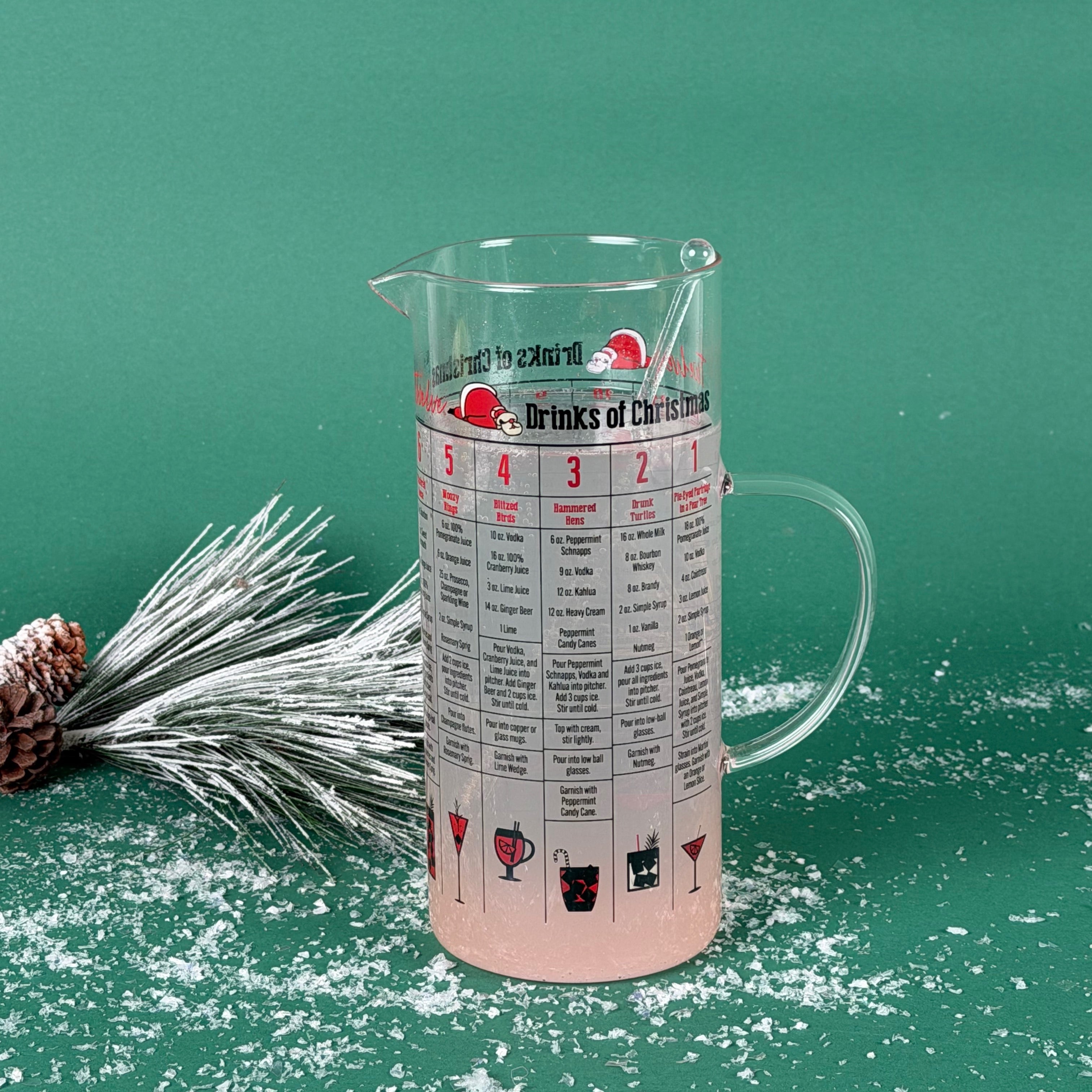 Clear Christmas-themed cocktail recipe pitcher filled with a light drink, shown on a snowy green background with frosted pine décor