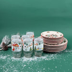 Christmas-themed drinking glasses and stacked holiday plates with winter village designs on a snowy green background