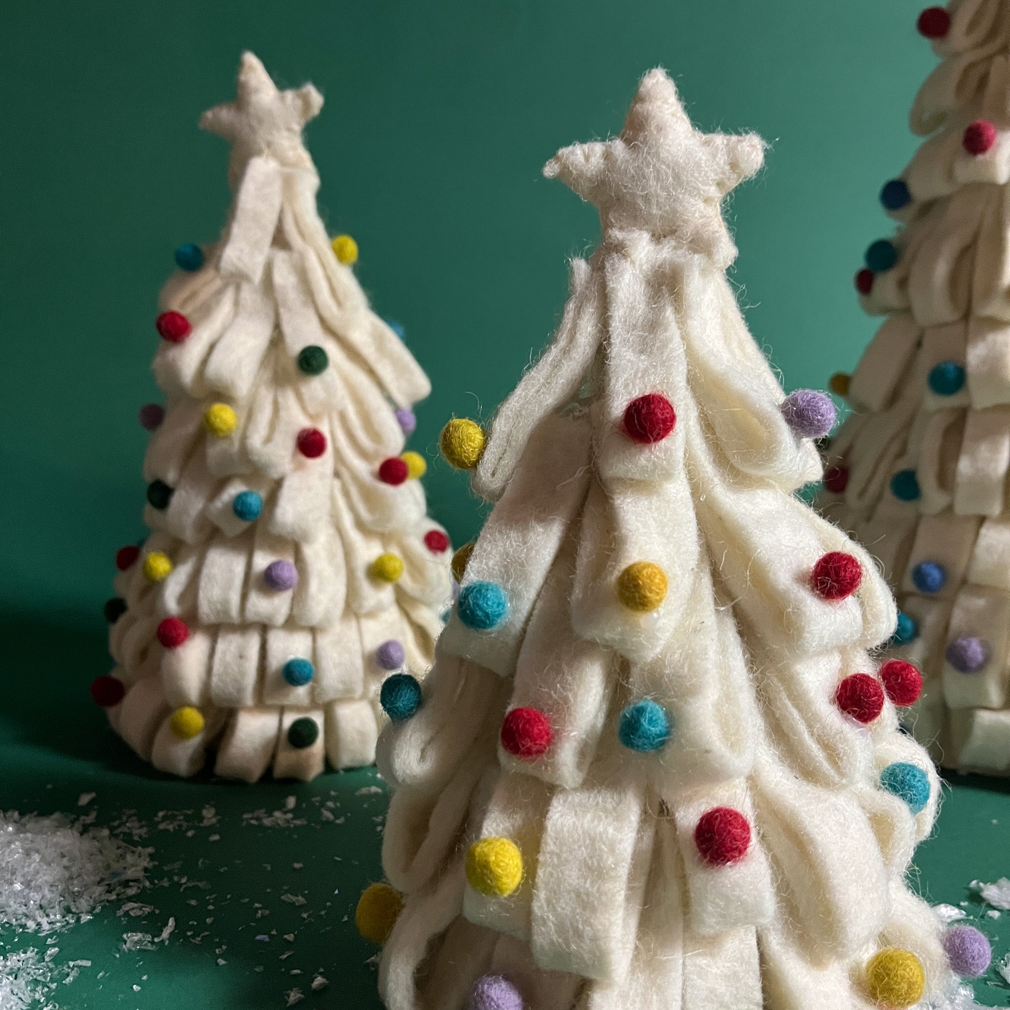 a set of three, white Looped Felt Christmas Tree Table Decorations with colourful bauble decorations, topped with stars against a green, snow scattered background. 