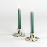 Two lit green taper candles displayed in white ceramic scalloped candle holders with hand-painted green brushstroke accents on a light background.