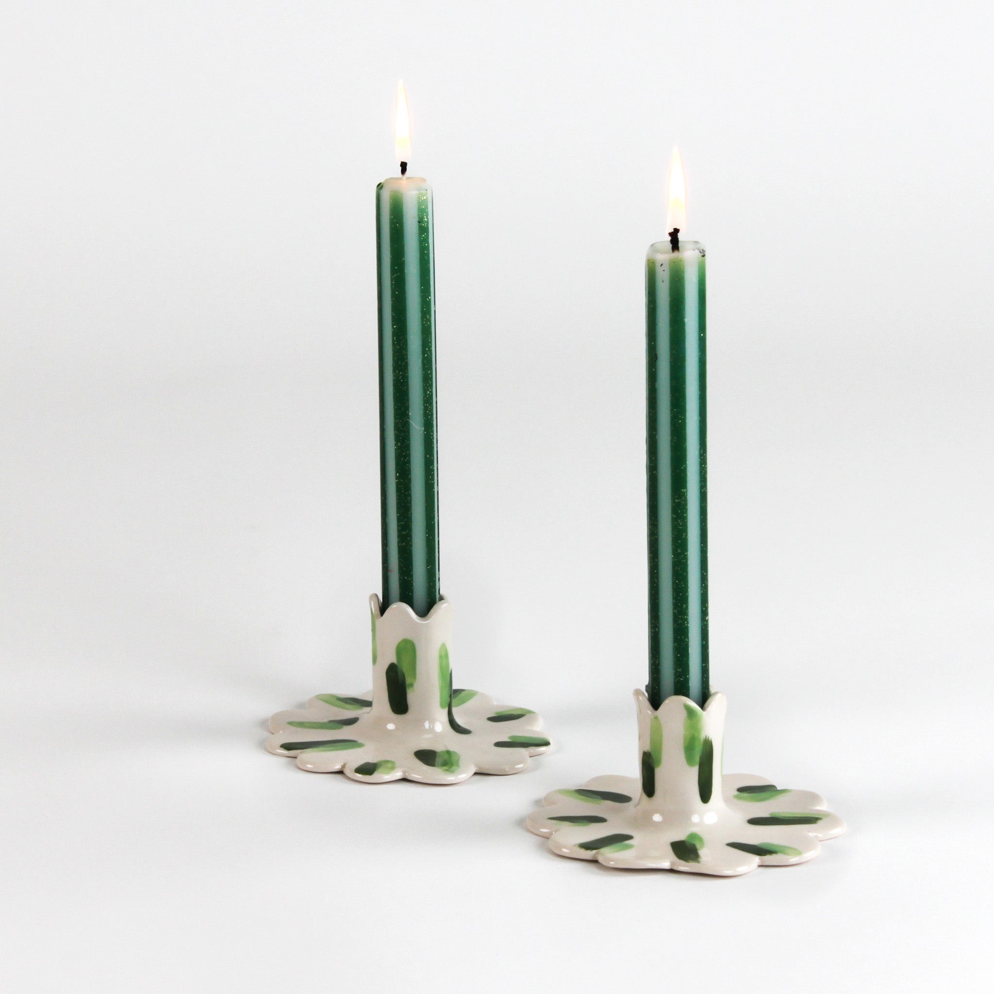 Two lit green taper candles displayed in white ceramic scalloped candle holders with hand-painted green brushstroke accents on a light background.
