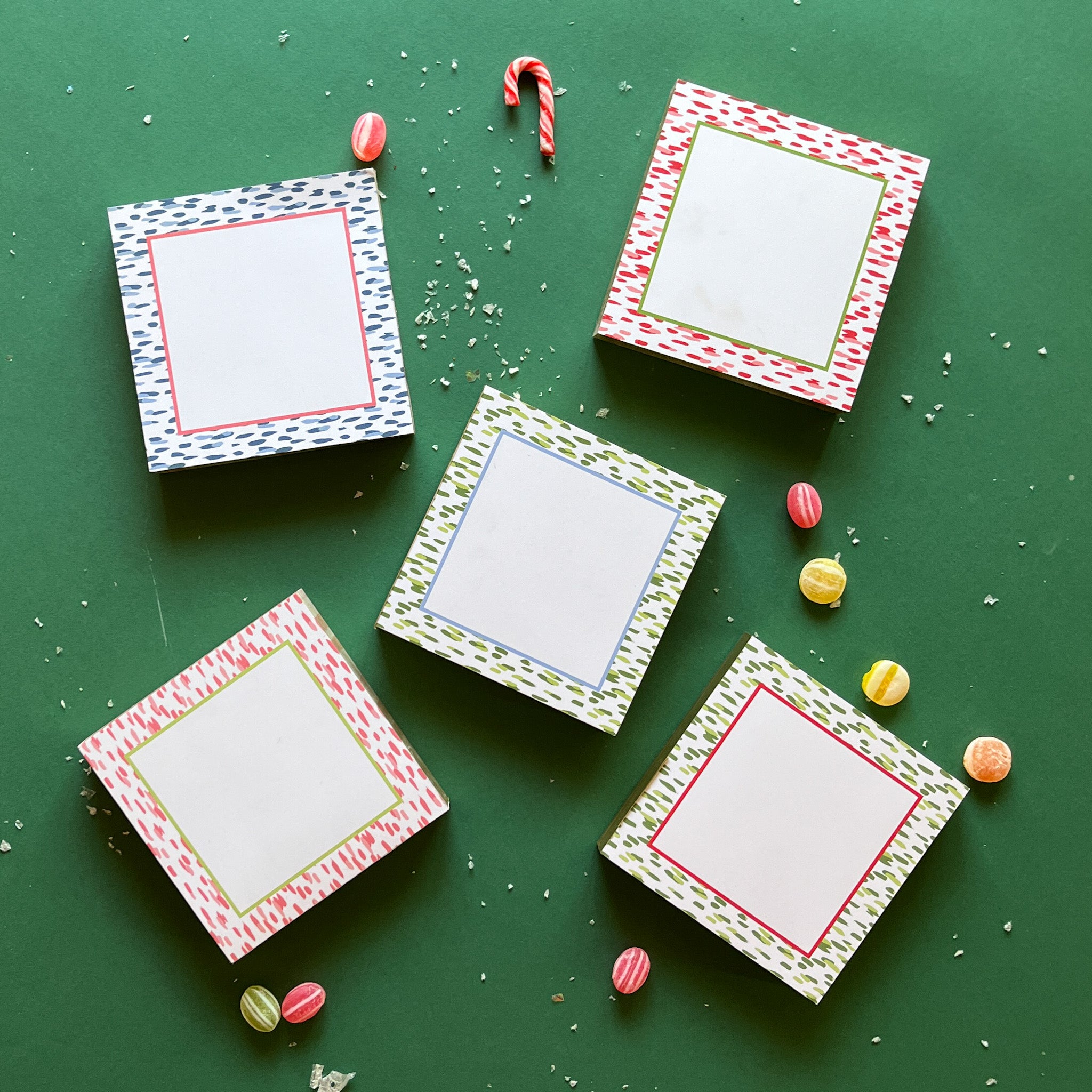 A collection of four square luxury notepads with colorful striped patterns on a green background, accompanied by candy canes and decorative candies.